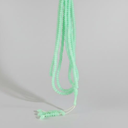 A hand holding a bundle of light green beaded strands, likely beads for jewelry making, against a busy background of other colorful beads and craft supplies.