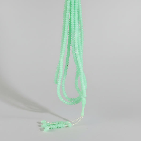 A hand holding a bundle of light green beaded strands, likely beads for jewelry making, against a busy background of other colorful beads and craft supplies.