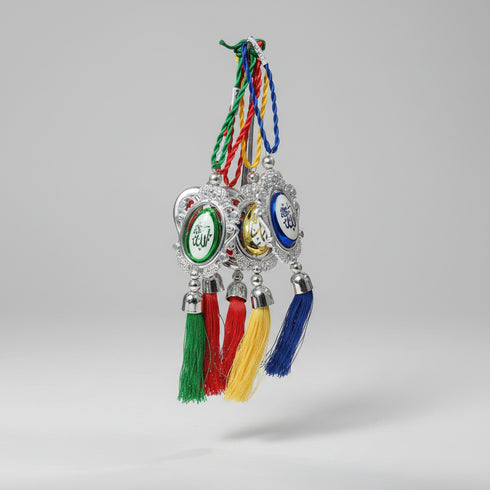 Three silver Islamic wall hanging ornaments with colorful tassels held in a person's hand. Each ornament features a different colored oval center with Arabic calligraphy: green, yellow, and blue, suspended from braided cords.