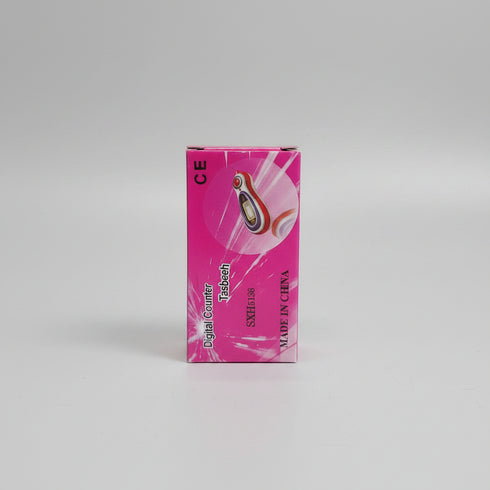 A hand holding a pink product box labeled 'Digital Counter Tasbeeh SXH5136' in an indoor setting with a cluttered desk in the background.