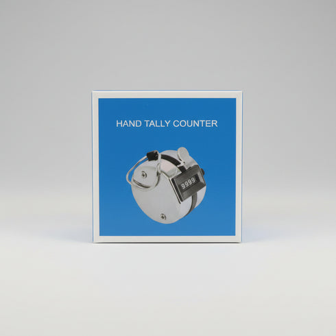 A hand holding a small square box labeled 'HAND TALLY COUNTER' with a blue front panel and a photo of a hand tally device. The background is a cluttered indoor desk or workshop setting.