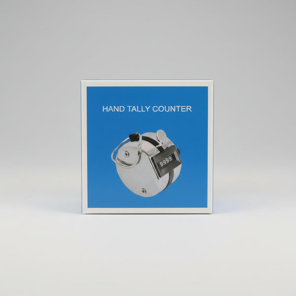 A hand holding a small square box labeled 'HAND TALLY COUNTER' with a blue front panel and a photo of a hand tally device. The background is a cluttered indoor desk or workshop setting.