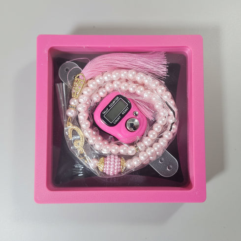 A pink gift box with a clear lid containing a pink digital fidget toy, a pink pearl beaded bracelet, and gold coins, held on a person's lap.