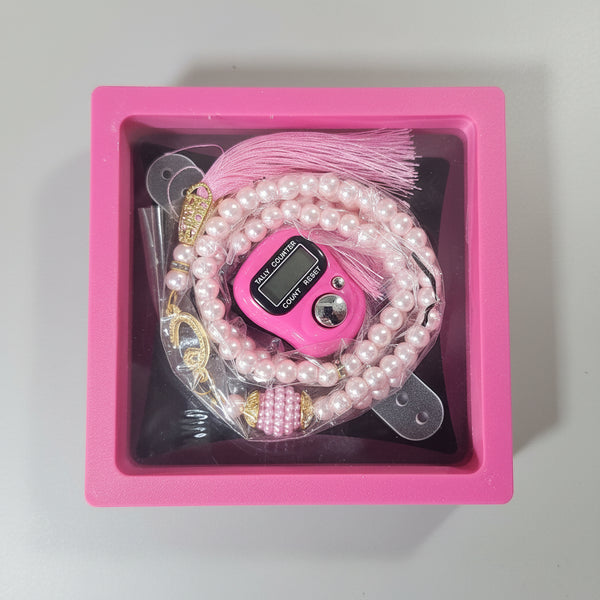 A pink gift box with a clear lid containing a pink digital fidget toy, a pink pearl beaded bracelet, and gold coins, held on a person's lap.
