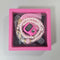 A pink gift box with a clear lid containing a pink digital fidget toy, a pink pearl beaded bracelet, and gold coins, held on a person's lap.
