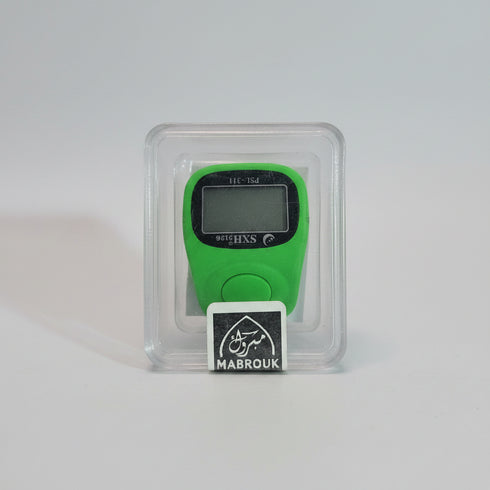 A green digital step counter inside a clear plastic clamshell package with a black and white MABROUK label, held by a hand against a blurred indoor background.