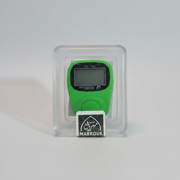A green digital step counter inside a clear plastic clamshell package with a black and white MABROUK label, held by a hand against a blurred indoor background.